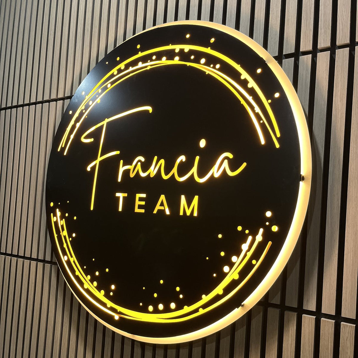 Close-up of the illuminated Francia Team sign showing glowing yellow script lettering and halo-lit edge on a wood slat wall